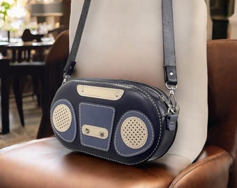 Könnte beinhalten: Eine schwarze Leder-Umhängetasche in Form eines Vintage-Radios. Die Tasche hat einen beigefarbenen Lederbesatz und zwei lautsprecherförmige Details mit einem perforierten Design.