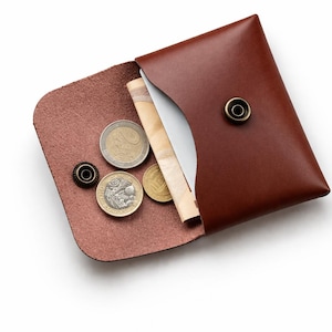 May include: A brown leather wallet, open to reveal coins and a folded bill. The wallet has a snap closure and a contrasting interior. The coins include a Euro coin, a British pound coin, and a smaller gold coin.