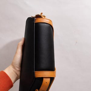 Peut inclure: Sac &agrave; main rectangulaire noir avec garniture en cuir beige et fermeture &agrave; boucle. Le sac a un design structur&eacute; avec une poign&eacute;e sup&eacute;rieure et une bandouli&egrave;re. Le sac est tenu par une personne.