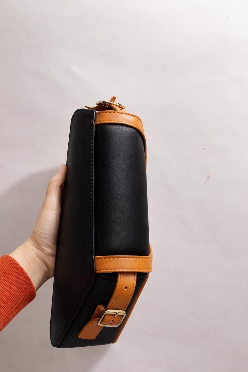 Peut inclure: Sac &agrave; main rectangulaire noir avec garniture en cuir beige et fermeture &agrave; boucle. Le sac a un design structur&eacute; avec une poign&eacute;e sup&eacute;rieure et une bandouli&egrave;re. Le sac est tenu par une personne.