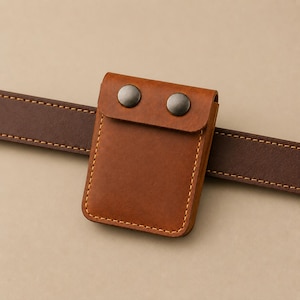 May include: A tan leather belt pouch featuring a rectangular shape with rounded corners, light-coloured stitching along the edges, and a flap secured by two dark metallic snap buttons, attached to a dark brown leather belt.