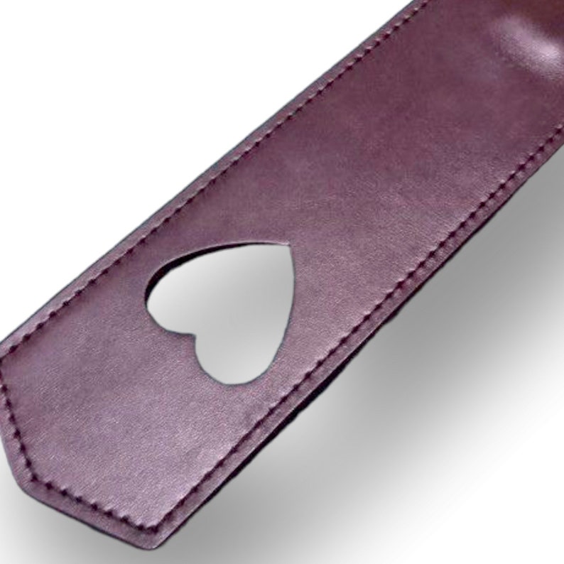 May include: A brown leather paddle with a heart-shaped cutout. The paddle has a stitched edge.