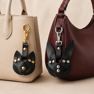Può includere: Due ciondoli per borsa in pelle nera a forma di testa di volpe, decorati con borchie metalliche. Uno ha finiture dorate attaccate a una borsa beige, l'altro ha finiture argentate attaccate a una borsa color bordeaux scuro.