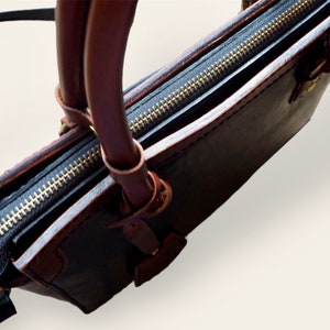 Pode incluir: Um close-up de uma bolsa de couro preta com al&ccedil;as e acabamentos em couro marrom. A bolsa apresenta um z&iacute;per dourado e um design estruturado. As al&ccedil;as s&atilde;o presas com an&eacute;is de metal.