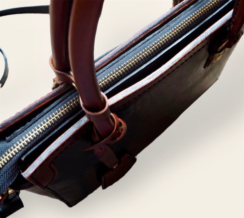 Pode incluir: Um close-up de uma bolsa de couro preta com al&ccedil;as e acabamentos em couro marrom. A bolsa apresenta um z&iacute;per dourado e um design estruturado. As al&ccedil;as s&atilde;o presas com an&eacute;is de metal.