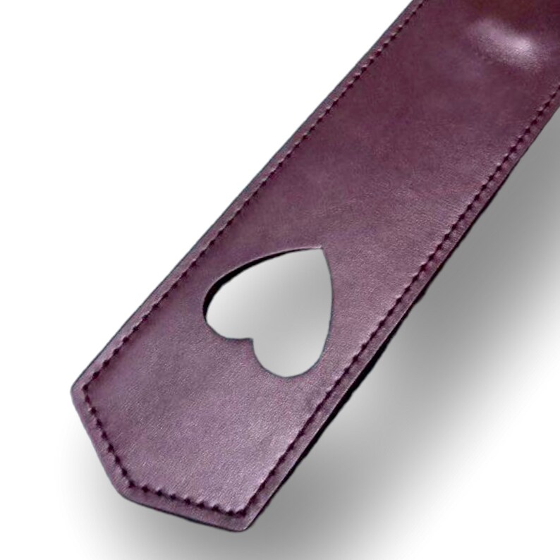 May include: A brown leather paddle with a heart-shaped cutout. The paddle has stitching around the edges.