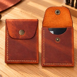 May include: Two rectangular brown leather card holders with visible light-colored stitching along the edges. One holder is closed with a brass snap button, while the other is open, revealing a black card inside and a matching brass snap closure.