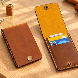 May include: Two vertical brown leather card holders with visible yellow stitching. One holder is closed with a brass snap button, while the other is open, revealing a yellow and blue card inside and a matching brass snap closure.