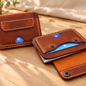 May include: Two brown leather card holders with visible stitching. One holder is closed, while the other is open, revealing a metal snap button closure and a slot containing a blue card and paper currency. Both items feature rounded corners.