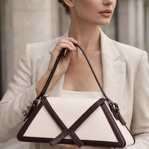 Peut inclure: Un sac &agrave; main cr&egrave;me avec un design g&eacute;om&eacute;trique. Le sac pr&eacute;sente un corps blanc avec des accents en cuir marron fonc&eacute; et une sangle assortie. Le sac est tenu par une personne portant un blazer beige.