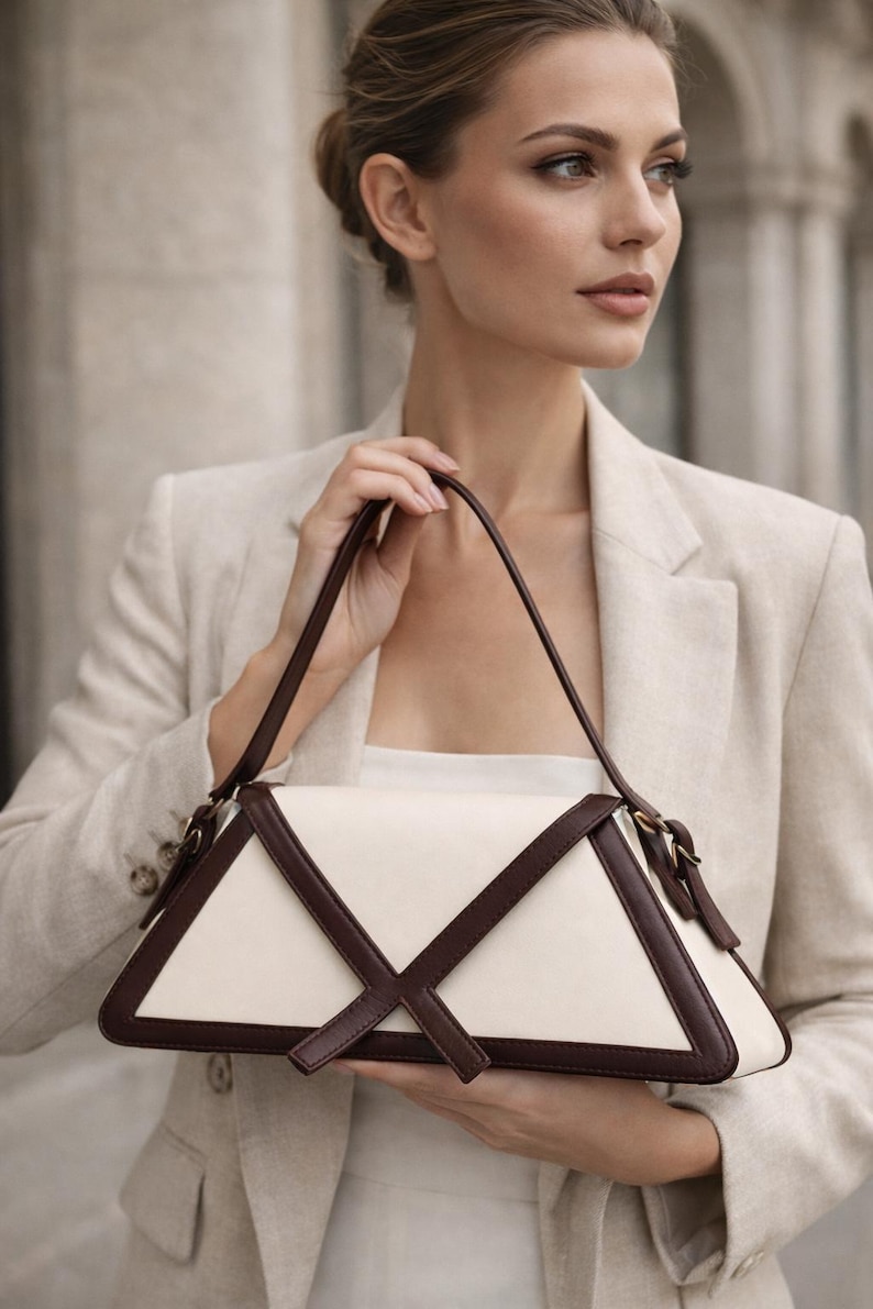 Peut inclure: Un sac &agrave; main cr&egrave;me avec un design g&eacute;om&eacute;trique. Le sac pr&eacute;sente un corps blanc avec des accents en cuir marron fonc&eacute; et une sangle assortie. Le sac est tenu par une personne portant un blazer beige.