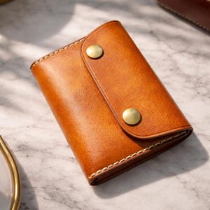 Puede incluir: Una pequeña cartera rectangular de cuero marrón con cierre de botón a presión. La cartera tiene una solapa asegurada por dos botones color latón. Los bordes están cosidos con hilo de color claro. La cartera está sobre una superficie de mármol.