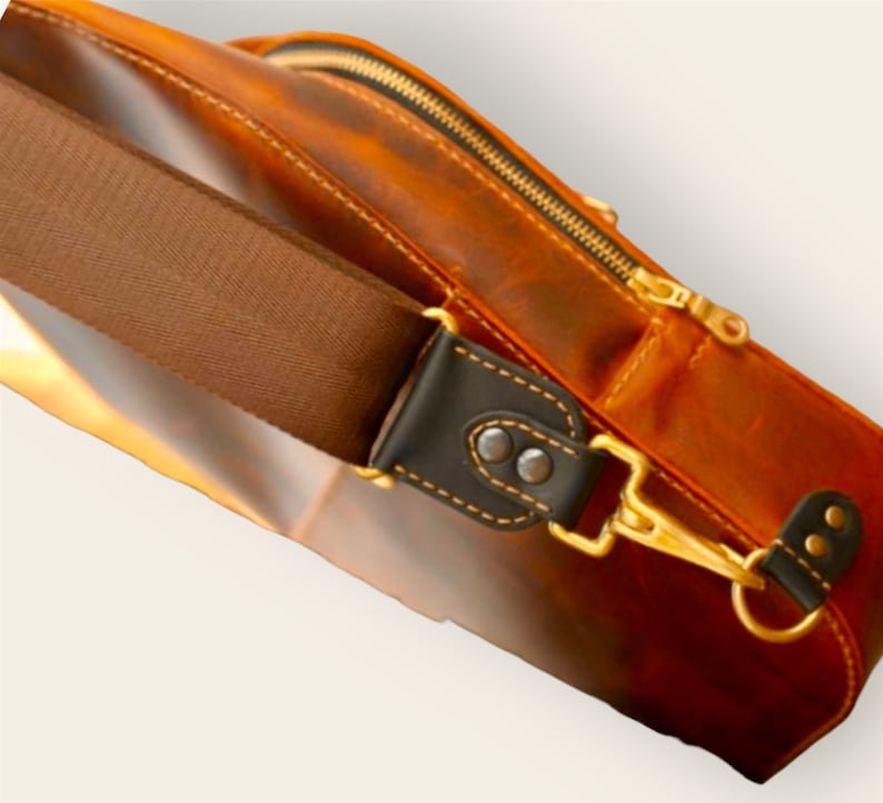 May include: A brown leather satchel bag with a zip closure and a brown adjustable strap. The strap has a gold metal clasp and a black leather accent.
