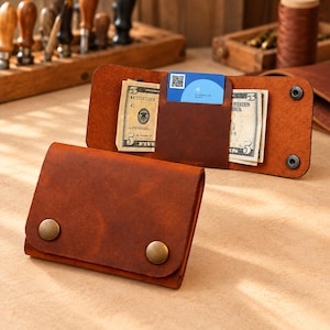 Puede incluir: Una cartera de cuero marrón con dos botones de presión de latón en la solapa frontal. El interior contiene dos billetes y una tarjeta azul con un código QR y el texto Istanbulkart, asegurados por un bolsillo central de cuero y broches.
