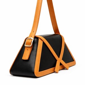 Peut inclure: Un sac &agrave; main noir de forme triangulaire unique, avec des accents en cuir de couleur tan. Le sac est dot&eacute; d'une poign&eacute;e et d'une bordure de couleur tan, avec une boucle dor&eacute;e. Le design comprend des lani&egrave;res de couleur tan qui se croisent sur le devant.
