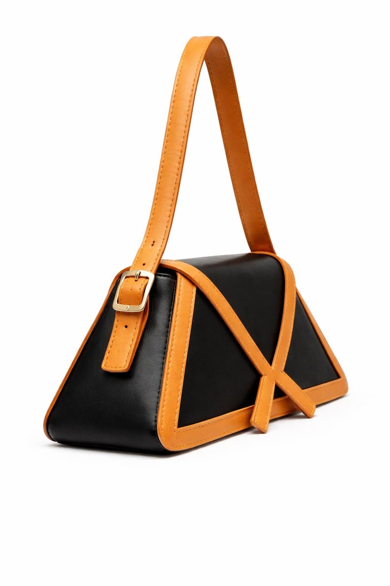 Peut inclure: Un sac &agrave; main noir de forme triangulaire unique, avec des accents en cuir de couleur tan. Le sac est dot&eacute; d'une poign&eacute;e et d'une bordure de couleur tan, avec une boucle dor&eacute;e. Le design comprend des lani&egrave;res de couleur tan qui se croisent sur le devant.