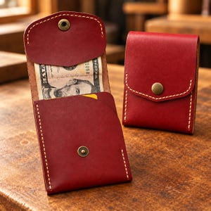 May include: Two red leather wallets with visible light-coloured stitching and antique brass snap closures. One wallet is open, revealing a five-dollar bill tucked inside, while the other wallet is closed and standing upright.