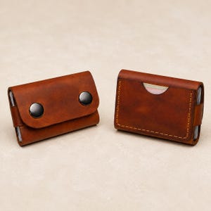 May include: Two brown leather card holders. The left holder features a flap secured by two dark metal snap buttons. The right holder has a rectangular design with visible stitching along the edges and a semi-circular cutout for card access.