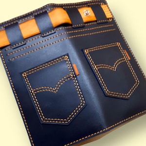May include: A blue leather wallet with a denim design. The wallet has two pockets with orange stitching and a brown leather strap with a button closure.