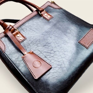 Pode incluir: Bolsa tote de couro preta e marrom com acabamento texturizado. A bolsa apresenta detalhes, al&ccedil;as e uma etiqueta remov&iacute;vel em couro marrom. Ferragens douradas adicionam detalhes. A bolsa tem formato retangular e uma al&ccedil;a de ombro.