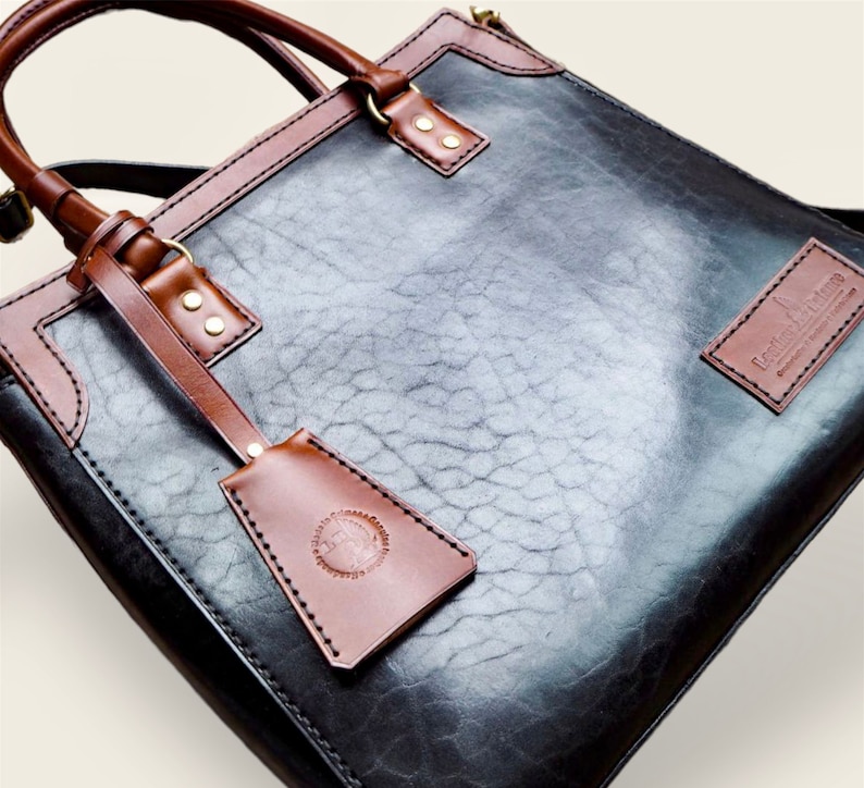 Pode incluir: Bolsa tote de couro preta e marrom com acabamento texturizado. A bolsa apresenta detalhes, al&ccedil;as e uma etiqueta remov&iacute;vel em couro marrom. Ferragens douradas adicionam detalhes. A bolsa tem formato retangular e uma al&ccedil;a de ombro.