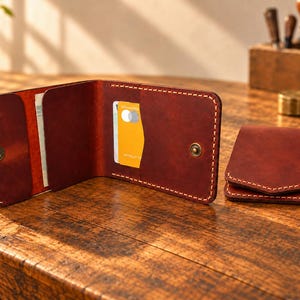 Può includere: Un portafoglio bifold in pelle rosso scuro con cuciture a vista di colore chiaro, dotato di chiusura a scatto in metallo, scomparti per carte e uno scomparto per banconote. Un secondo portafoglio piegato si trova a destra di quello aperto.