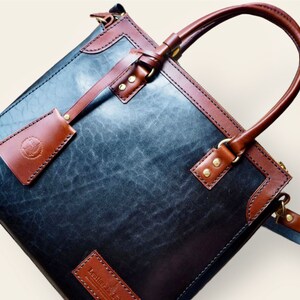 Pode incluir: Bolsa de couro artesanal com corpo azul-cinzento escuro e acabamentos castanhos. Possui uma al&ccedil;a superior, uma etiqueta remov&iacute;vel e ferragens de lat&atilde;o. O couro tem uma apar&ecirc;ncia texturizada. A bolsa parece nova.