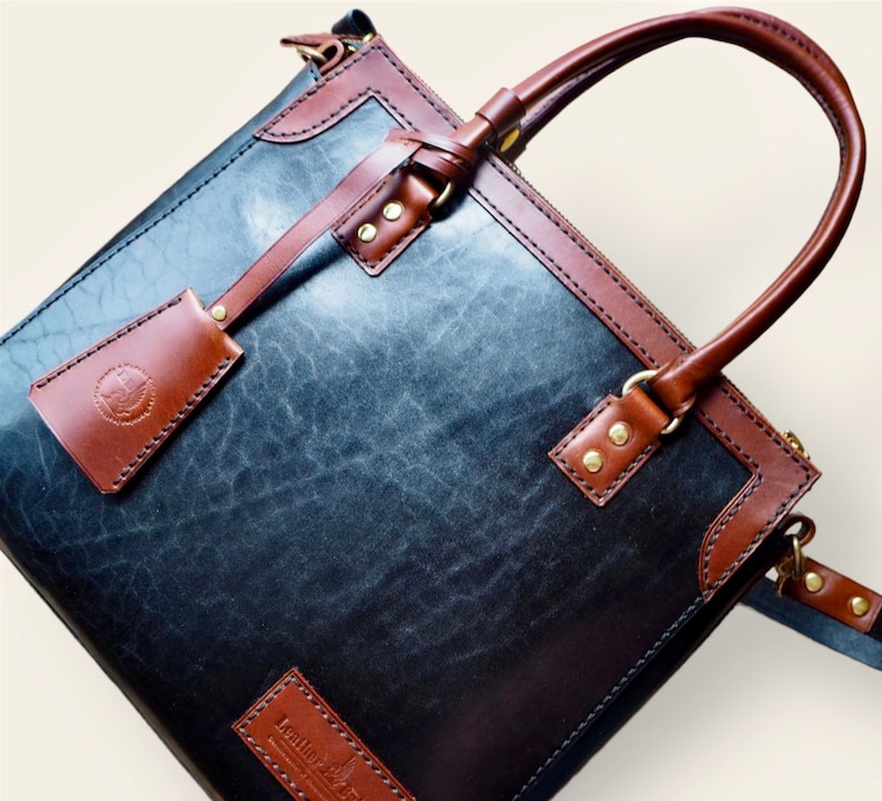 Pode incluir: Bolsa de couro artesanal com corpo azul-cinzento escuro e acabamentos castanhos. Possui uma al&ccedil;a superior, uma etiqueta remov&iacute;vel e ferragens de lat&atilde;o. O couro tem uma apar&ecirc;ncia texturizada. A bolsa parece nova.