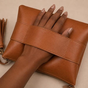 May include: A tan leather clutch bag featuring a wide horizontal strap across the front, a zipped closure, and a matching leather tassel attached to a side ring. The rectangular bag has a smooth finish and clean, minimalist stitching throughout.