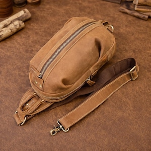 Op de afbeelding: Een bruine leren sling bag met een hoofdvak met ritssluiting en een verstelbare schouderriem. De tas heeft een ruige, versleten uitstraling, met zichtbare stiksels en metalen hardware. De riem is bevestigd met een metalen sluiting.