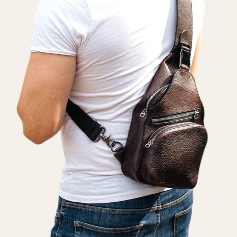 Peut inclure: Un sac &agrave; bandouli&egrave;re en cuir marron avec plusieurs compartiments &agrave; fermeture &eacute;clair. Le sac est port&eacute; sur l'&eacute;paule d'une personne portant un t-shirt blanc et un jean bleu.