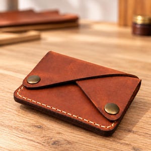 May include: A brown leather card holder with a folded envelope-style flap secured by two antique brass-colored metal snap buttons. The edges are stitched with light-colored thread, and the leather has a smooth, matte finish.
