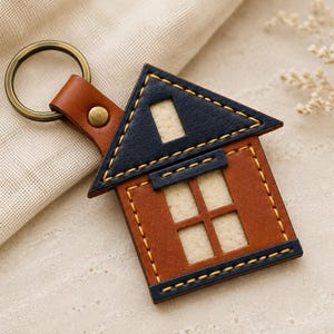Pode incluir: Um porta-chaves de couro em forma de casa com corpo em couro castanho, telhado e base em couro azul escuro. O design inclui uma janela recortada e um espaço de sótão, acentuado com costuras amarelas e um anel de metal em tom de bronze.