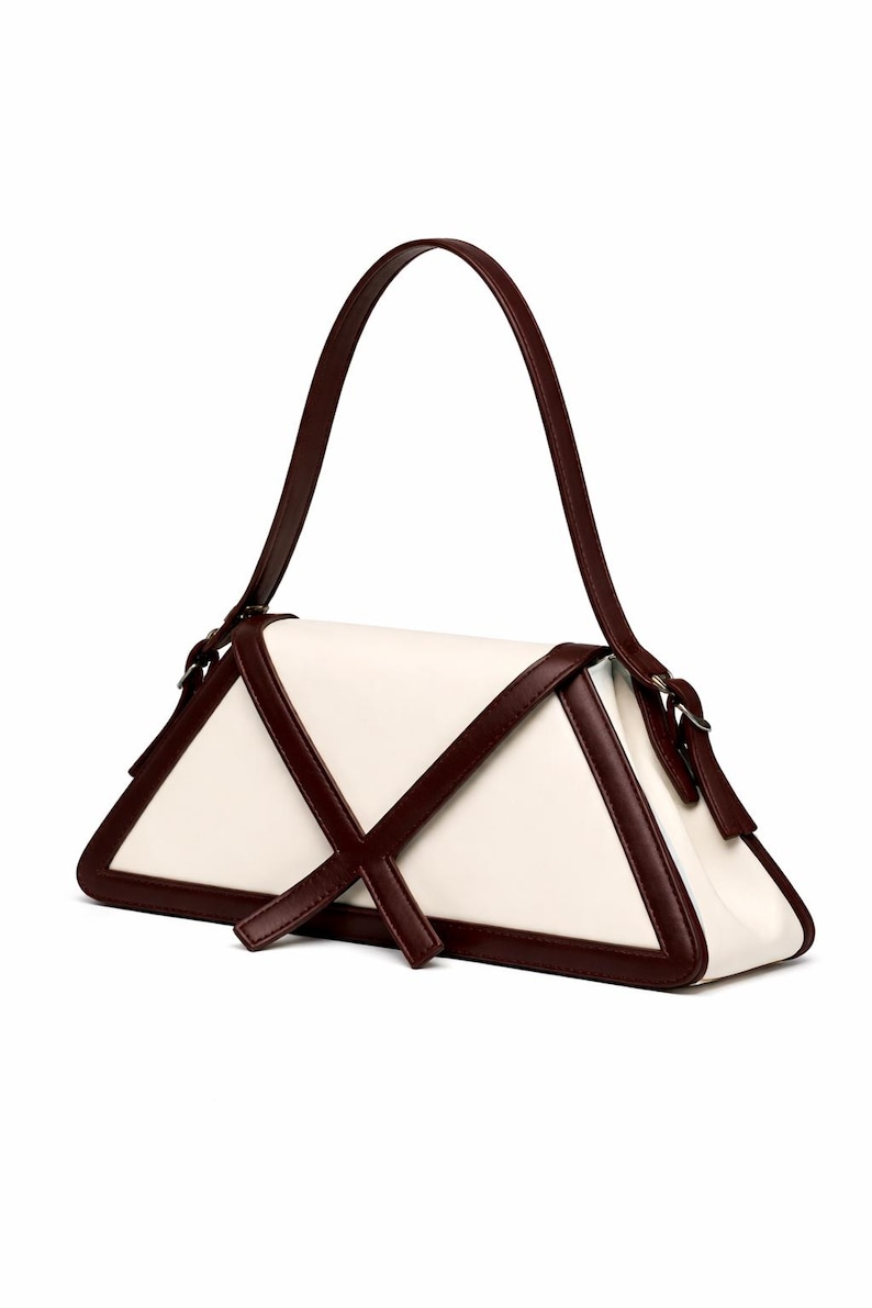 Peut inclure: Sac &agrave; main g&eacute;om&eacute;trique de forme triangulaire. Le corps est de couleur cr&egrave;me, avec des bordures et des accents marron fonc&eacute;. Il poss&egrave;de une poign&eacute;e unique et un motif en forme de croix sur le devant.