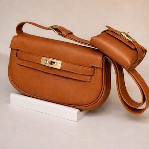 May include: A brown leather handbag with a matching small pouch. The handbag has a gold-colored clasp and a long strap. The pouch is attached to the strap. The bag is sitting on a white surface.