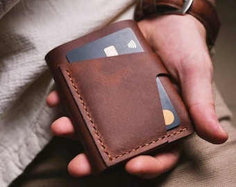 May include: A brown leather wallet with a card slot and a flap closure. The wallet is held in a hand.