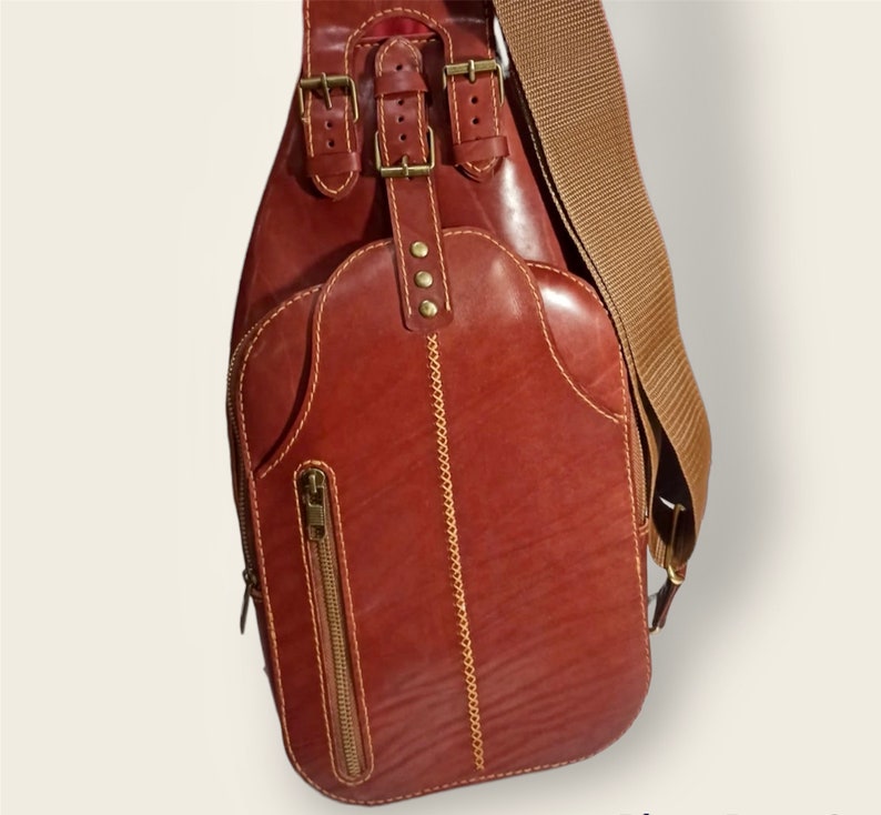 May include: A brown leather crossbody bag with a long adjustable strap. The bag has a flap closure with multiple buckles and a zipped pocket on the front.