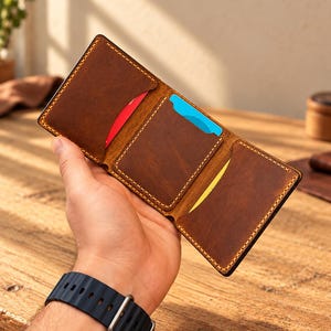 Puede incluir: Una cartera de cuero marrón de tres pliegues con costuras claras visibles en los bordes. La cartera tiene tres ranuras interiores para tarjetas, cada una con una tarjeta con un borde superior rojo, azul o amarillo visible.