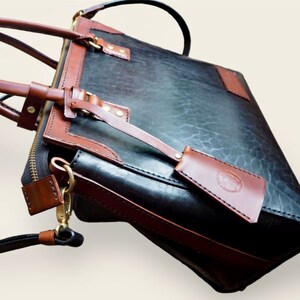 Pode incluir: Uma pasta de couro preto e castanho com uma al&ccedil;a superior e uma al&ccedil;a de ombro remov&iacute;vel. A bolsa possui um bolso frontal, um fecho de z&iacute;per e detalhes em couro castanho. A bolsa tem uma etiqueta de bagagem anexada.