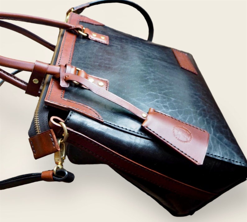 Pode incluir: Uma pasta de couro preto e castanho com uma al&ccedil;a superior e uma al&ccedil;a de ombro remov&iacute;vel. A bolsa possui um bolso frontal, um fecho de z&iacute;per e detalhes em couro castanho. A bolsa tem uma etiqueta de bagagem anexada.
