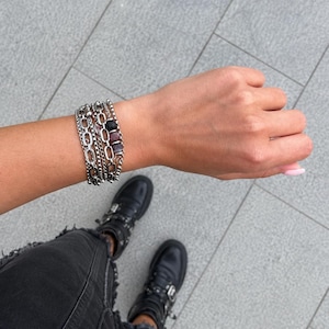 May include: A multi-strand silver-toned bracelet featuring a combination of thick oval links, delicate cable chains, and a series of small beads in black, dark purple, and light pink, worn on a wrist with black jeans and black leather boots.