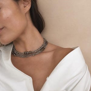 May include: A multi-layered silver-toned chain necklace featuring a combination of delicate link chains and small, dark, spherical beads. The necklace rests against the neck, paired with a crisp, white collared shirt worn off the shoulder.