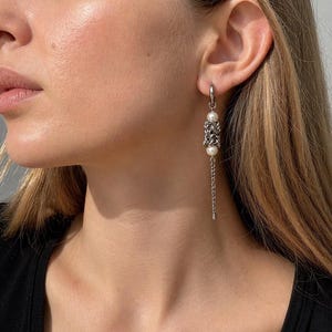 Pode incluir: Brincos pendurados prateados com um design de malha de corrente e duas pérolas brancas. Os brincos pendem de um pequeno aro e apresentam uma corrente delicada. A joia é mostrada na orelha de uma pessoa.