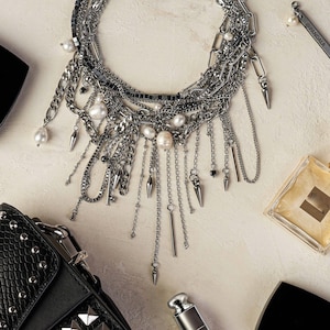 Puede incluir: Un collar de cadenas plateadas de varias capas con diversos estilos de eslabones, perlas blancas y puntas metálicas colgantes. Incluye un pequeño dije rectangular con el texto PANDALA, un bolso negro con tachuelas, perfume y brillo labial.