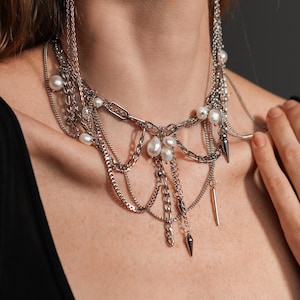 Peut inclure: Un collier à chaînes en métal argenté superposées avec plusieurs rangs drapés, agrémenté de perles d'eau douce blanches et de petites pointes métalliques. Le design inclut un long segment de chaîne relié à une boucle d'oreille assortie.