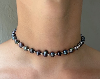 May include: A choker necklace featuring a single strand of dark, iridescent, baroque-shaped pearls interspersed with small, round, polished silver-toned metal beads. The necklace sits closely around the neck, showcasing the varied shapes and dark hues.