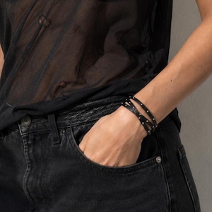 Op de afbeelding: Een zwarte, meerstrengs leren armband met kleine zilverkleurige metalen studs, gedragen om de pols. De armband bestaat uit verschillende dunne, overlappende bandjes die om de arm wikkelen, wat zorgt voor een gelaagde uitstraling met metalen accenten.
