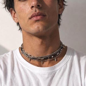 May include: A layered silver-tone chain necklace featuring a combination of a thick paperclip chain, a delicate rope chain, and a dark beaded strand. The necklace is accented with multiple small, pointed silver-tone spikes hanging from the chains.