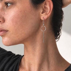 Pode incluir: Um brinco pendente de tom prateado com uma pequena conta de metal presa a um gancho fino, seguida por um anel circular e um pingente de barra vertical fino. O brinco pende do lóbulo da orelha, exibindo um design metálico e geométrico.