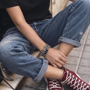 Puede incluir: Una camiseta negra, pantalones vaqueros azules con dobladillos y zapatillas altas de color burdeos con cordones blancos. Una pulsera de cadena plateada de varias capas se lleva en la muñeca. El conjunto tiene un estilo casual con detalles desgastados.